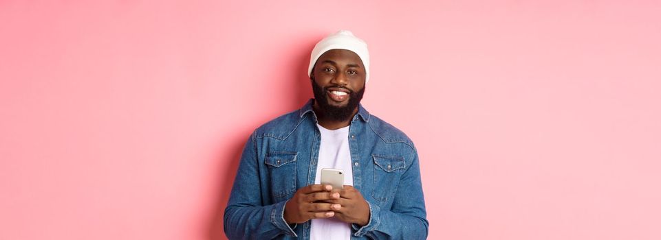 Handsome african-american guy using smartphone, smiling at camera, standing over pink background.