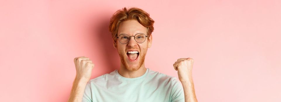 Close up of satisfied lucky redhead man winning, shouting from joy and making fist pump, celebratig victory, standing like champion over pink background.