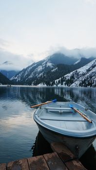 A mountain lake in the forest with mirrored water. Wooden pier with white boats. The water reflects the landscape of a cloudy sky, snowy mountains and peaks, coniferous firs. Kolsai Lake, Kazakhstan