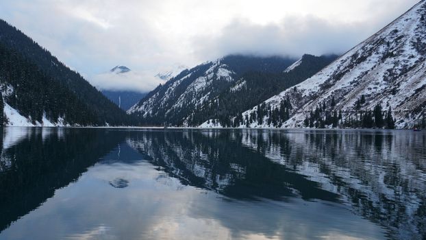 Kolsai mountain lake in the winter forest. Drone view of clouds, coniferous trees, mirrored smooth water, hills and mountains in the snow. Yellow sunset. Boats float in places. Kazakhstan, Almaty