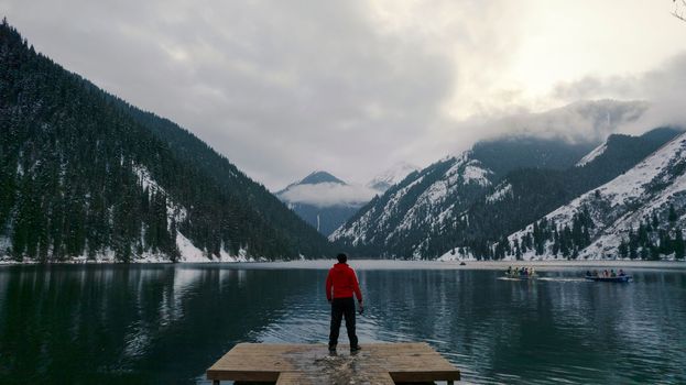 The guy standing on the pier admires the mountain lake. There is a view of the mirrored black color of the water, which reflects snowy mountains, green forest, clouds and a yellow sunset. Kolsai Lake