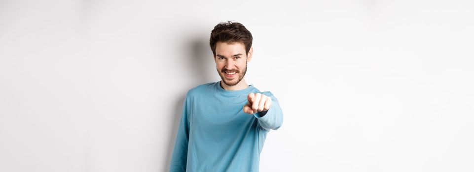 Image of confident young man smiling and pointing at camera, inviting and choosing you, beckon to join him, standing on white background.