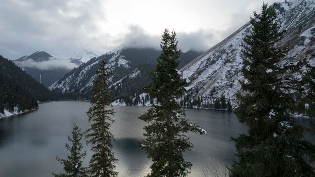 Kolsai mountain lake in the winter forest. Drone view of coniferous trees, mirror-smooth water like a mirror, hills, mountains in snow and clouds. Yellow sunset. Boats float in places. Kazakhstan