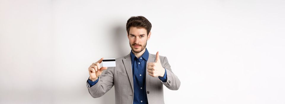 Very good bank. Businessman in suit show plastic credit card and thumb-up, recommending, standing on white background.