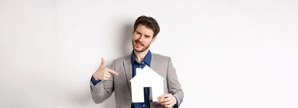 Real estate and insurance concept. Salesman in grey suit showing paper house cutout, selling property, smiling at camera, white background.