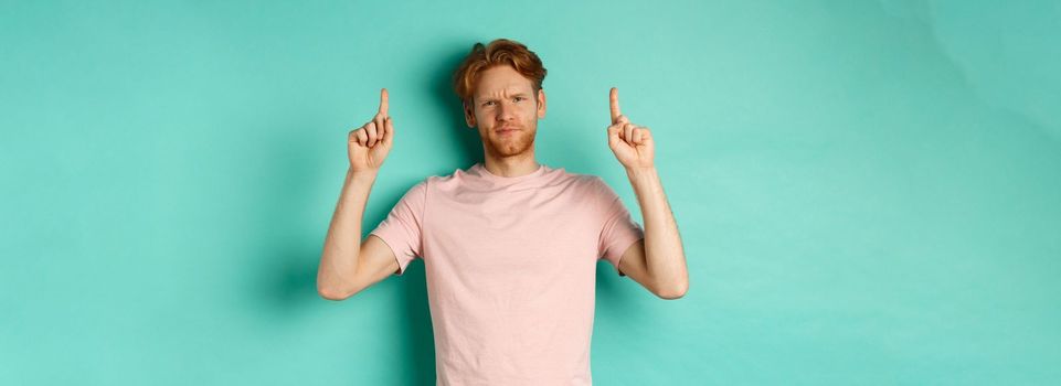 Skeptical and disappointed redhead man pointing fingers up, frowning displeased and looking upset, standing over turquoise background.