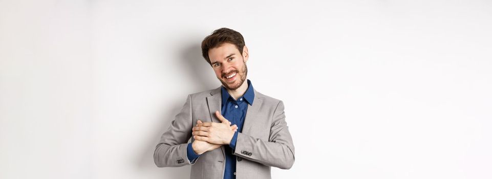 Happy man in suit holding hands on heart, feeling touched or grateful, having tender memories in soul, standing against white background.