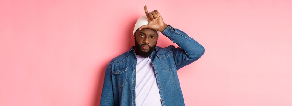 Sad african-american man showing loser sign on forehead and staring at camera, standing over pink background.