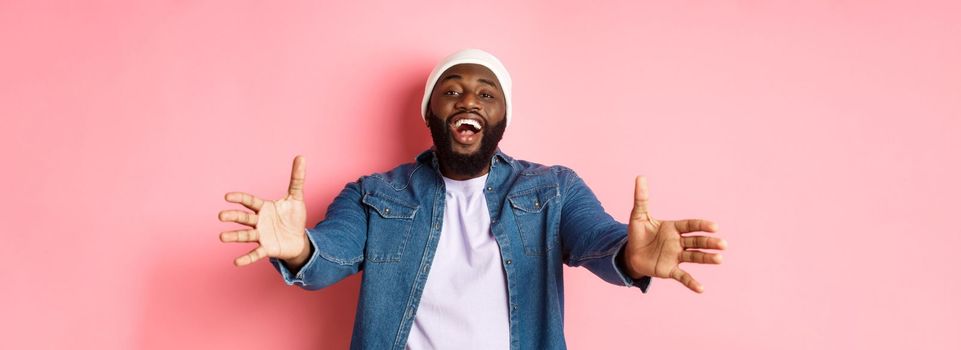 Happy Black man stretching hands, reaching for hug or take something, smiling pleased, standing in beanie and denim shirt over pink background.