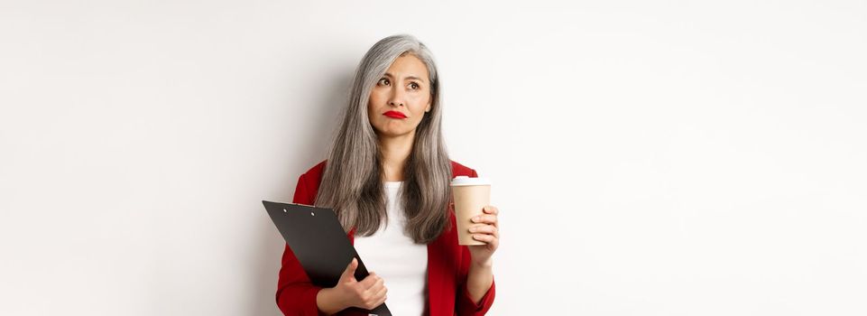 Sad asian businesswoman drinking coffee at work and looking upper left corner, standing over white background.