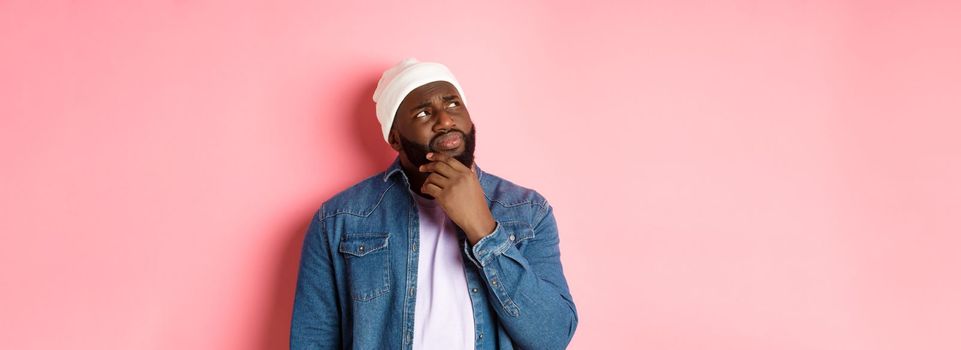 Image of puzzled and concerned black man, staring at upper left corner and thinking, making decision with doubts, standing over pink background.