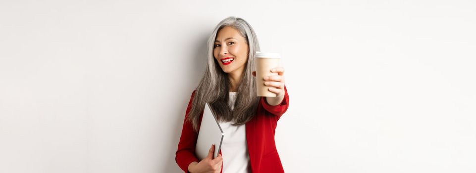 Business. Cheerful asian woman manager giving you cup of coffee and smiling, standing with laptop in hand, white background.