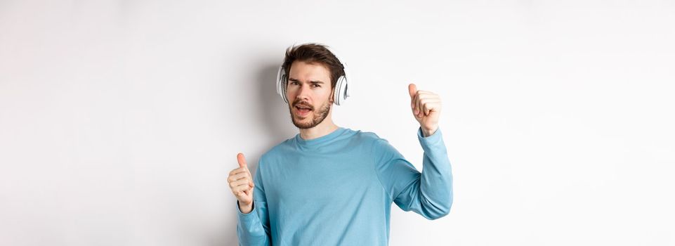 Cheeky guy dancing and having fun in wireless headphones, enjoying good sound quality, white background.