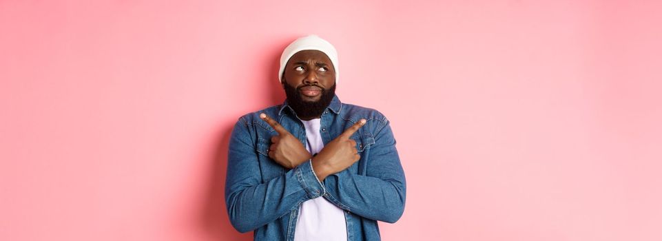 Confused Black man with beard, making choice, pointing fingers sideways and looking puzzled, standing over pink background.