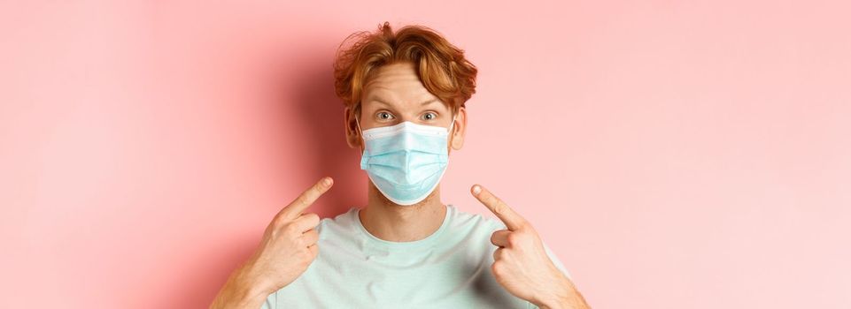 Covid-19 and pandemic concept. Close up of redhead man with messy hairstyle, pointing fingers at medical mask from coronavirus, standing over pink background.