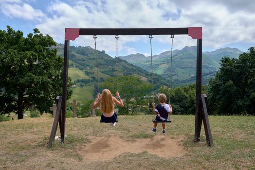 Woman and blonde child on a swing. Caucasian, unrecognisable, mountain, wooden swing, lifestyle, mountains with meadows, summer, sunny and clouds