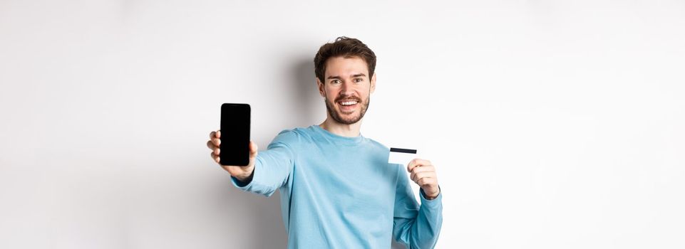 E-commerce and shopping concept. Smiling caucasian man showing plastic credit card and empty smartphone screen, recommending online app, white background.
