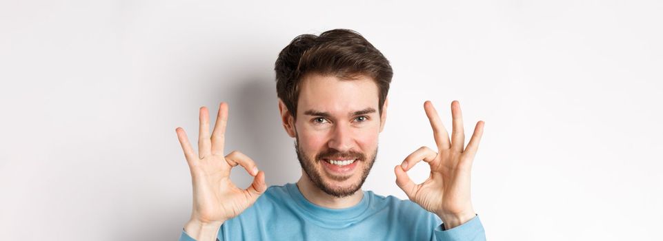 Close-up of confident young man smiling, showing okay signs, praising excellent choice, approve something good, standing over white background.