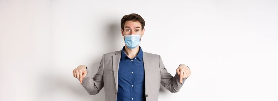 Covid-19, pandemic and business concept. Surprised male ceo manager in medical mask and suit pointing fingers down, looking amused, white background.