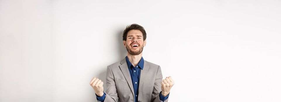 Excited man winning in cazino and celebrating, making fist pump and shouting yes with happy expression, achieve goal, triumphing, standing on white background.