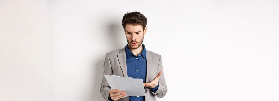 Confused businessman looking at bad document, complaining on deal, pointing at paper frustrated, standing in suit on white background.