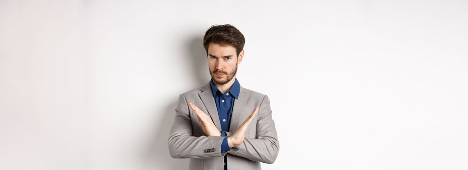 Serious business man making cross and says no, looking determined, disagree and prohibit something bad, tell to stop, standing in suit on white background.