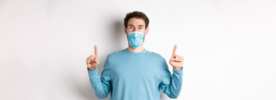 Coronavirus, health and quarantine concept. Excited smiling guy checking out special deal, pointing fingers up, stare amazed at camera, wear medical mask on white background.