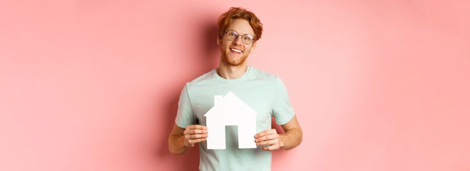 Real estate. Cheerful young man with red hair, wearing glasses and t-shirt, showing paper house cutout and smiling, buying apartment, pink background.