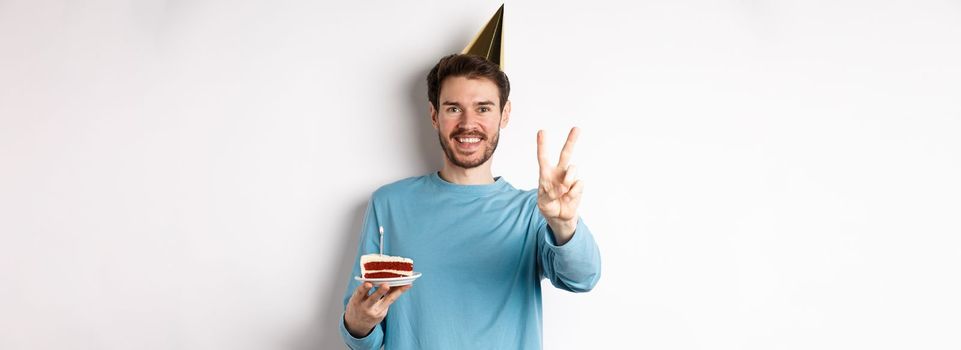 Celebration and holidays concept. Happy young man celebrating birthday, taking picture with peace sign, wearing party hat and holding bday cake, white background.