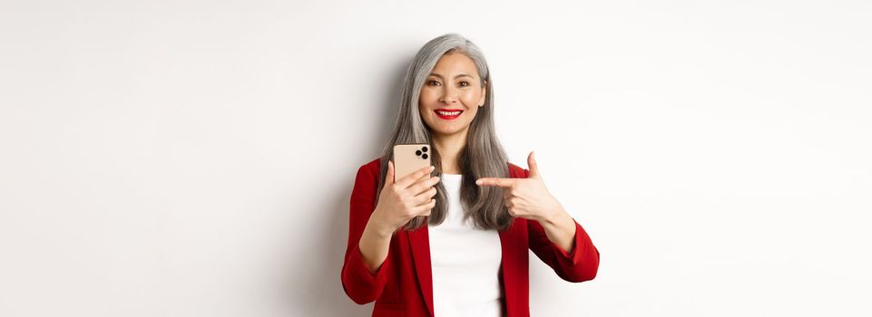 Asian elderly woman in elegant blazer showing smartphone, pointing finger at mobile phone and smiling, standing over white background.