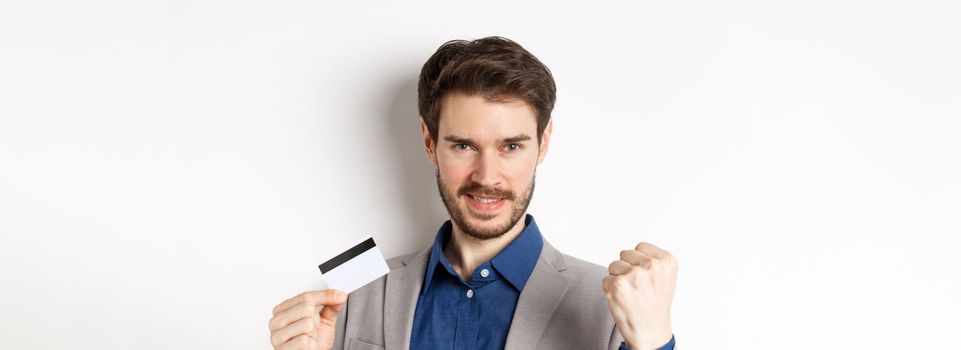 Shopping. Cheerful handsome businessman showing plastic credit card and fist pump, saying yes with rejoice, making money, standing on white background.