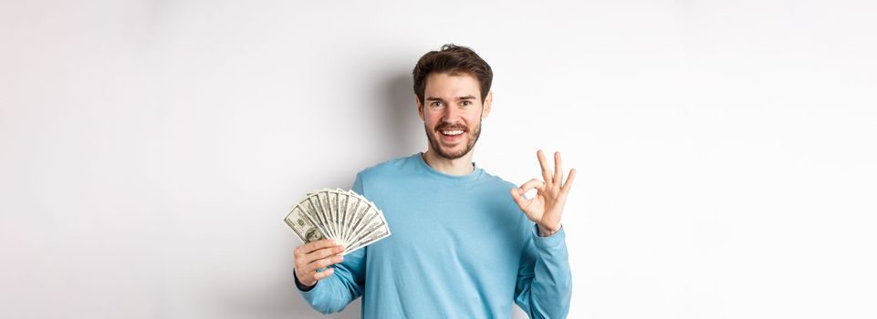Handsome young man showing quick loans money, make okay gesture and smiling with cash, standing over white background.