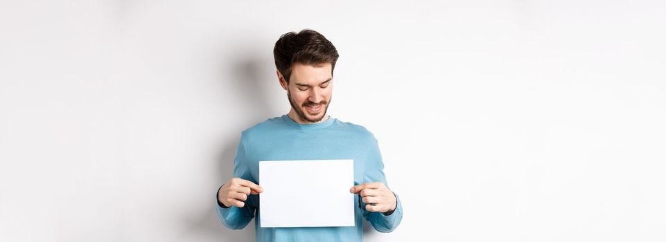 Young caucasian man looking at empty paper, reading logo, standing on white background.