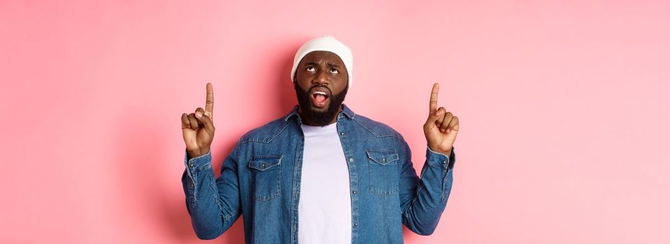 Displeased african american guy in beanie, pointing fingers up and staring at camera bothered, complaining, standing over pink background.