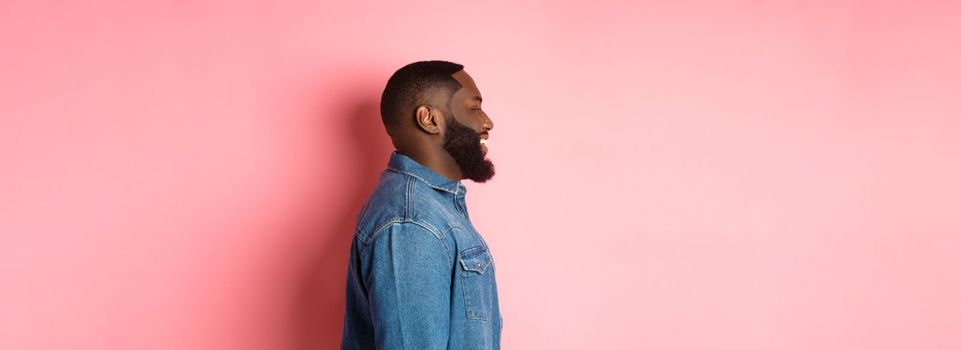Profile of handsome bearded Black guy standing over pink background, smiling and looking left at copy space.