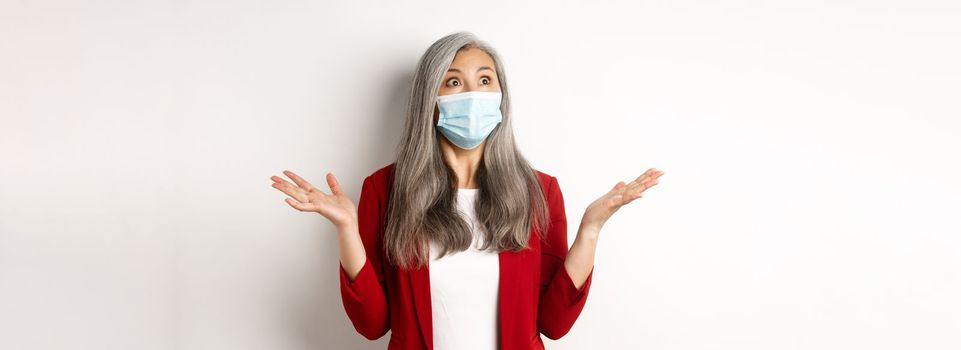 Coronavirus and business concept. Surprised asian businesswoman in face mask, spread hands sideways and staring shocked at upper left corner, white background.