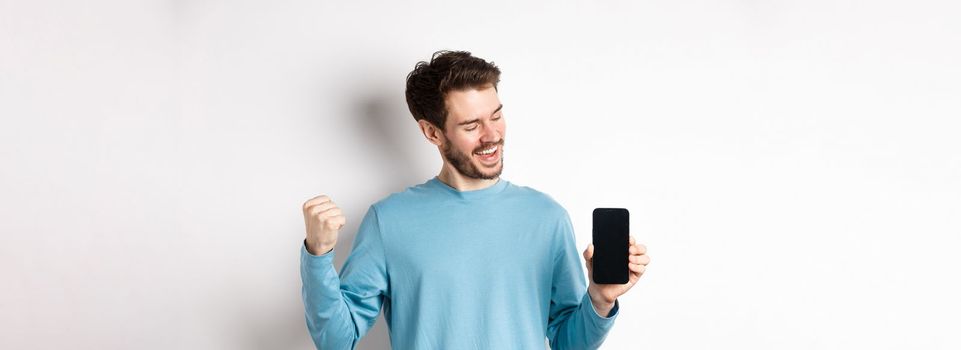 E-commerce and shopping concept. Lucky man winning online, showing empty smartphone screen and triumphing, saying yes with fist pump, standing over white background.