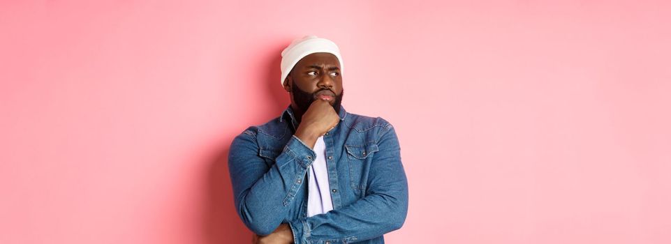 Serious thoughtful african american male model making decision, looking left concerned, thinking or having doubts, standing over pink background.
