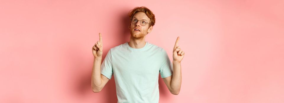 Dreamy young man with red hair and beard pointing fingers up, looking pensive at top, standing against pink background.