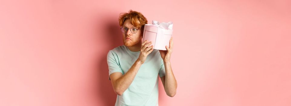 Love and holidays concept. Intrigued redhead guy press ear to box and shaking gift, guessing what inside, standing over pink background.
