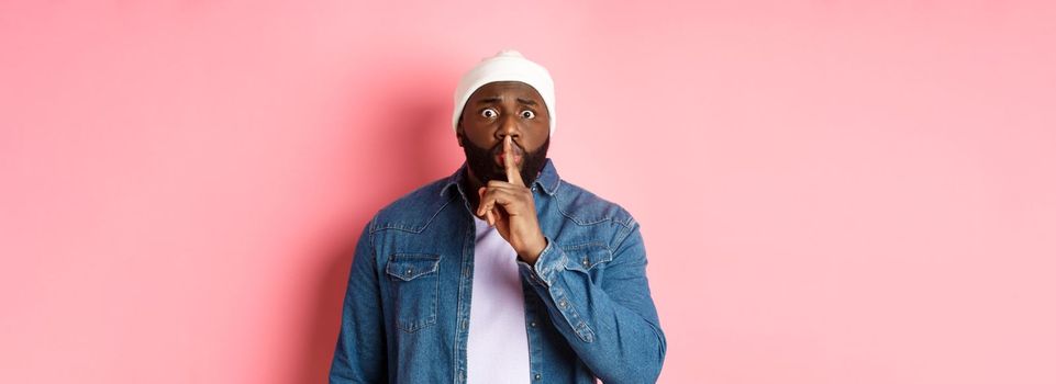 Worried Black man asking to keep quiet, sharing secret and hushing at you, holding finger pressed to lips and staring nervously at camera, pink background.