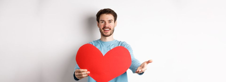 Handsome man in love making confession to you, pointing hand at camera, holding big red heart cutout on valentines day, singing romantic serenade, standing over white background.