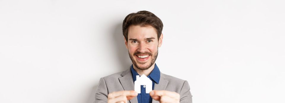 Real estate and insurance concept. Handsome man in suit smiling, showing small paper house cutout, standing on white background.