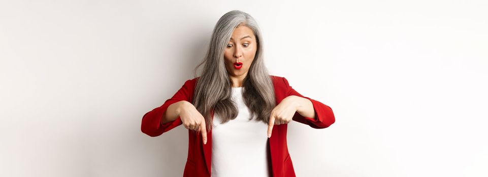 Impressed asian mature woman checking out promo, pointing and looking down with amazed face, standing in blazer over white background.