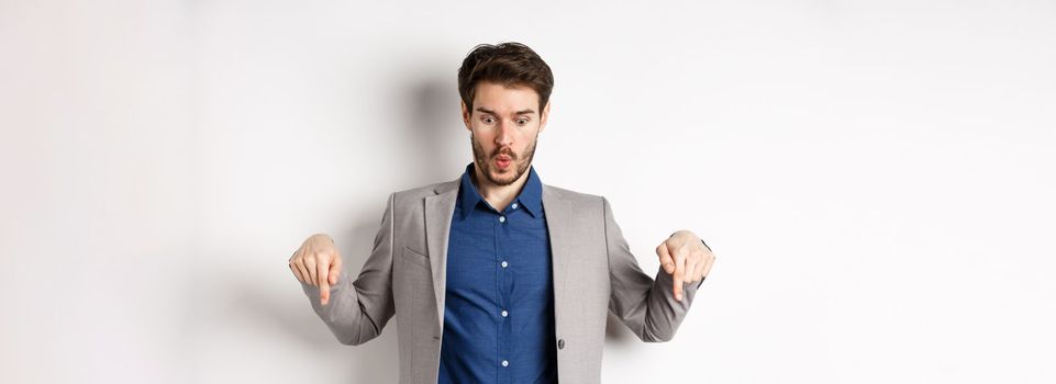 Wow look at this. Impressed and excited businessman in suit pointing fingers down, looking at bottom advertisement with amazed face, standing on white background.