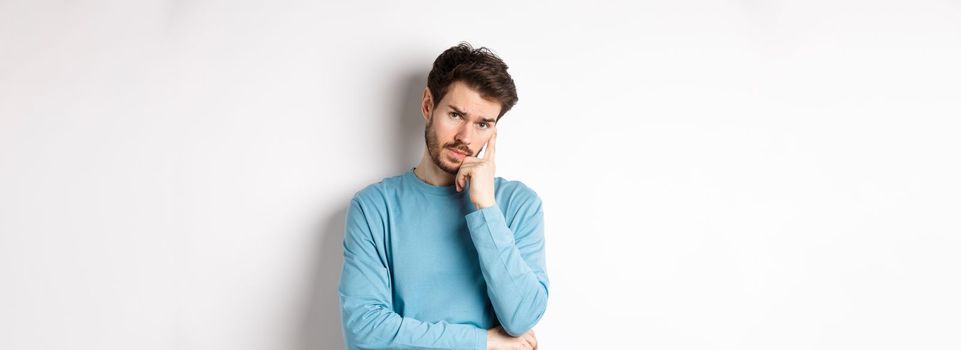 Skeptical young man listening to you with bored face, looking at camera reluctant, standing in sweatshirt over white background.