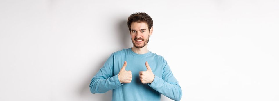 Good choice. Attractive caucasian man with beard showing thumbs up and smiling, like and approve good product, recommending something, standing on white background.