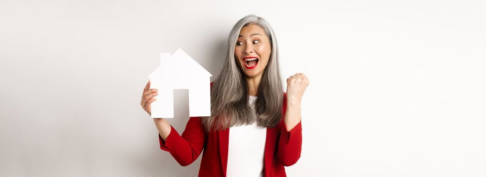 Cheerful asian senior woman buying house, scream of joy and making fist pump while showing paper house cutout, standing over white background.