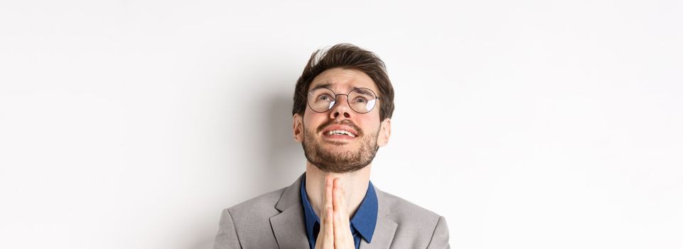 Nervous hopeful man in glasses and suit begging god, asking please and shaking hands in pray, white background.