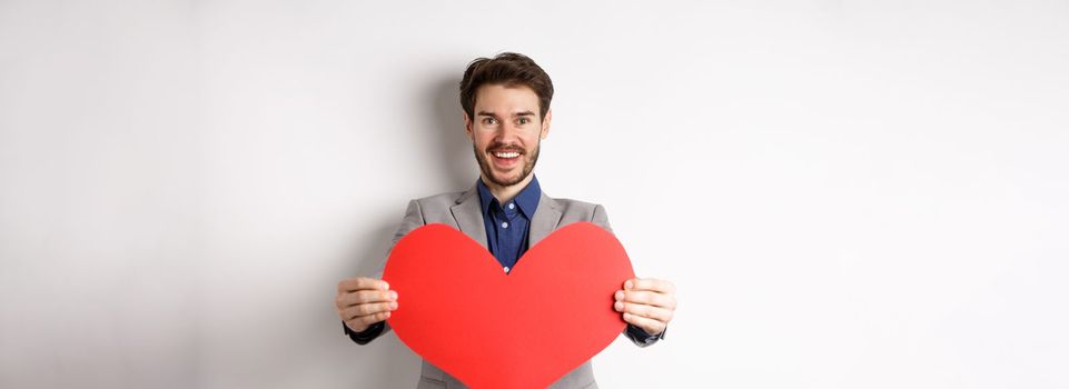 Handsome young man wishing happy Valentines day, giving big red heart sign and smiling, make surprise to lover, standing in suit over white background.